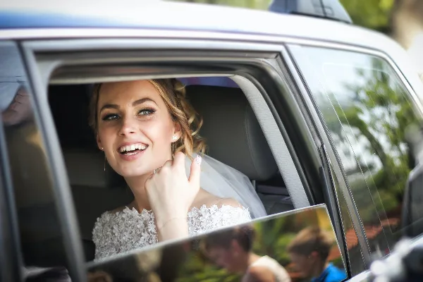 A smiling bride in a white wedding dress sitting in the back seat of a car.