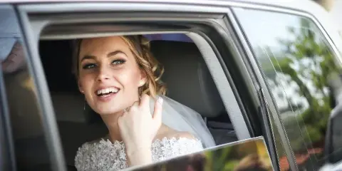 A smiling bride in a white wedding dress sitting in the back seat of a car.