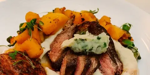 Plated dish of sliced roast beef over mashed potatoes with roasted potatoes and herb garnish on a white plate.