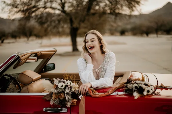 A smiling bride in a white dress sitting in a red convertible car decorated with flowers.