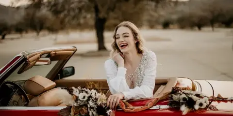 A smiling bride in a white dress sitting in a red convertible car decorated with flowers.