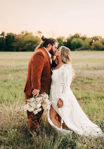 A bride and groom kissing in a grassy field at sunset, with the groom wearing a rust-colored suit and the bride in a white lace dress holding a bouquet.