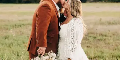 A bride and groom kissing in a grassy field at sunset, with the groom wearing a rust-colored suit and the bride in a white lace dress holding a bouquet.