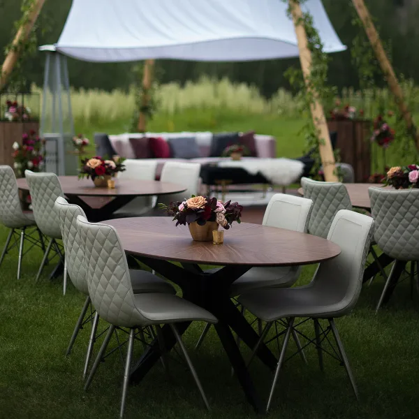 An outdoor event setup featuring round wooden tables, modern chairs, floral centerpieces, and a lounge area with a sofa and canopy.