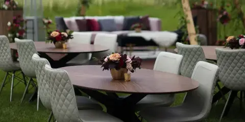 An outdoor event setup featuring round wooden tables, modern chairs, floral centerpieces, and a lounge area with a sofa and canopy.