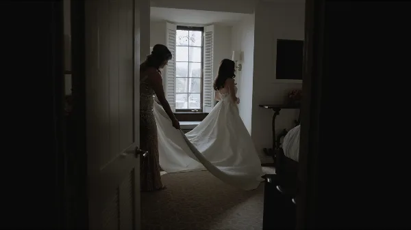 A bride and her mother in a hotel room, with the mother holding the train of the bride's white wedding dress.