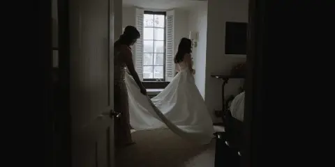 A bride and her mother in a hotel room, with the mother holding the train of the bride's white wedding dress.