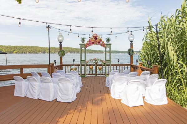 A wedding ceremony setup with white chairs and a floral arch on a wooden deck overlooking a lake.