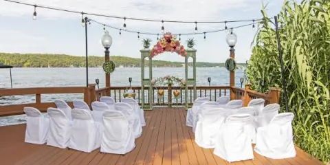 A wedding ceremony setup with white chairs and a floral arch on a wooden deck overlooking a lake.