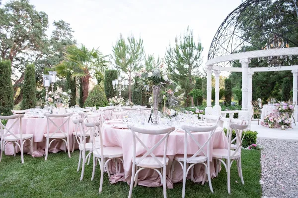 An outdoor wedding reception setup with white cross-back chairs, tables draped in pink linens, and floral centerpieces.