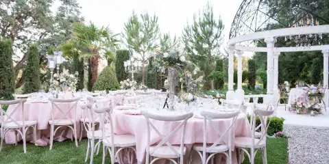 An outdoor wedding reception setup with white cross-back chairs, tables draped in pink linens, and floral centerpieces.