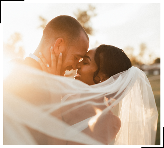 A close-up, artistic wedding photograph of a couple sharing an intimate moment, with the bride's veil draped over them in soft, golden light.