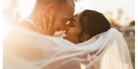 A close-up, artistic wedding photograph of a couple sharing an intimate moment, with the bride's veil draped over them in soft, golden light.