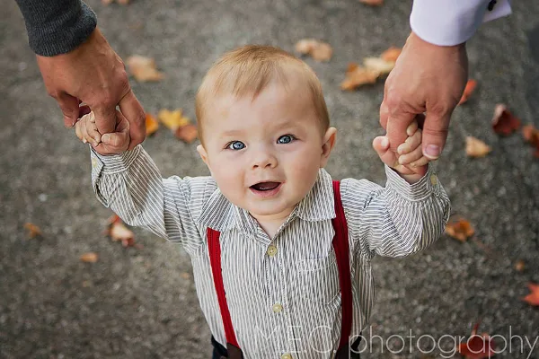 A smiling toddler in a striped shirt and red suspenders is being held by the hands of an adult, with autumn leaves on the ground in the background.