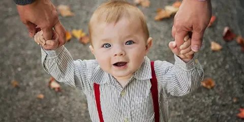 A smiling toddler in a striped shirt and red suspenders is being held by the hands of an adult, with autumn leaves on the ground in the background.
