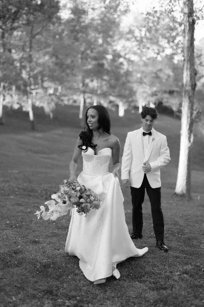 Black-and-white photograph of a bride in a white gown holding a bouquet walking outdoors with a groom in a white jacket.