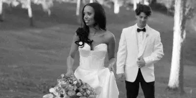 Black-and-white photograph of a bride in a white gown holding a bouquet walking outdoors with a groom in a white jacket.