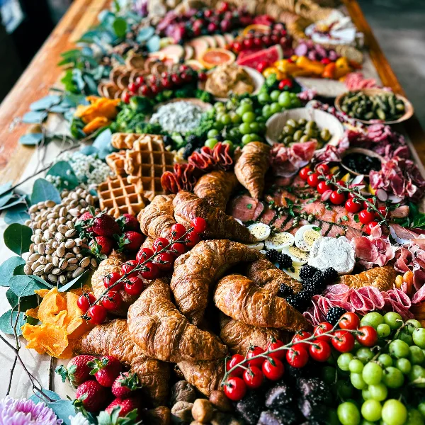 A large, colorful catering spread on a wooden table featuring croissants, cured meats, cheeses, grapes, and strawberries.