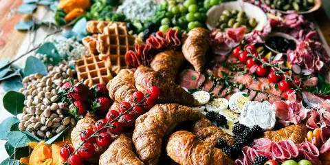 A large, colorful catering spread on a wooden table featuring croissants, cured meats, cheeses, grapes, and strawberries.