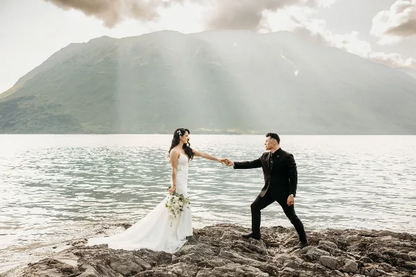 A bride and groom holding hands on a rocky shoreline with a large mountain in the background.