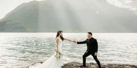 A bride and groom holding hands on a rocky shoreline with a large mountain in the background.