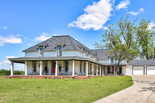 A large white farmhouse with a covered porch, manicured lawn, and driveway under a blue sky.