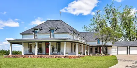 A large white farmhouse with a covered porch, manicured lawn, and driveway under a blue sky.