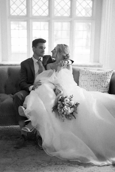 Black and white photograph of a bride and groom sitting together on a sofa, with the bride holding a bouquet.