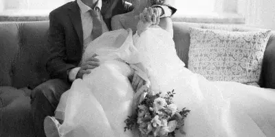 Black and white photograph of a bride and groom sitting together on a sofa, with the bride holding a bouquet.