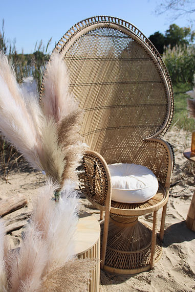 A tall woven wicker chair with a white cushion and pampas grass arrangement styled on a sandy outdoor setting.