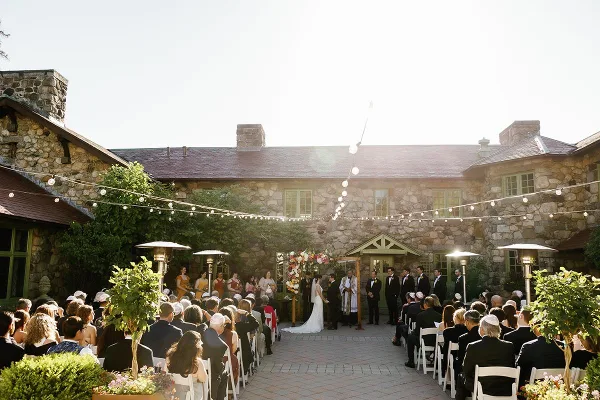 Outdoor wedding ceremony with guests seated in white chairs, string lights overhead, and a stone mansion in the background.