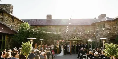Outdoor wedding ceremony with guests seated in white chairs, string lights overhead, and a stone mansion in the background.
