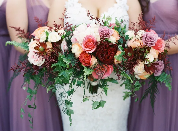 A bride in a white lace dress holding a large bouquet of pink, peach, and burgundy roses and dahlias, flanked by bridesmaids in purple dresses holding matching bouquets.