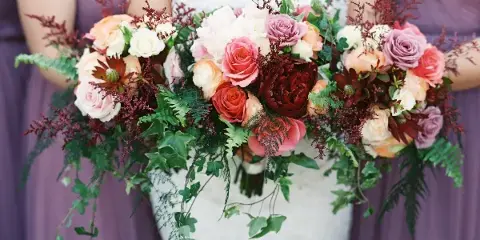 A bride in a white lace dress holding a large bouquet of pink, peach, and burgundy roses and dahlias, flanked by bridesmaids in purple dresses holding matching bouquets.