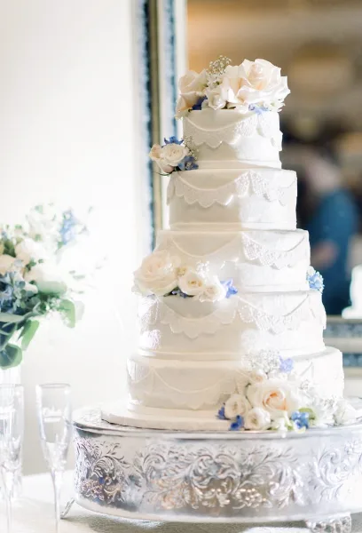 A five-tier white wedding cake decorated with white roses and blue accents, displayed on a silver stand.