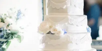 A five-tier white wedding cake decorated with white roses and blue accents, displayed on a silver stand.