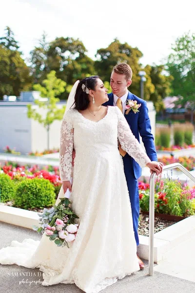 A bride in a white lace gown and a groom in a blue suit standing together outdoors in a garden setting.