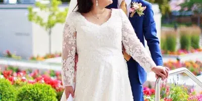 A bride in a white lace gown and a groom in a blue suit standing together outdoors in a garden setting.