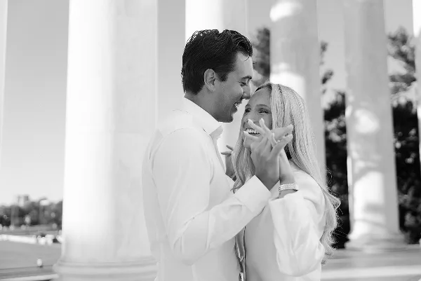 A black and white photograph of a smiling couple laughing together in front of classical columns, likely at the Thomas Jefferson Memorial.