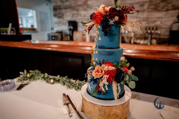 A three-tier blue wedding cake decorated with fresh flowers and gold accents on a table with a cake server.