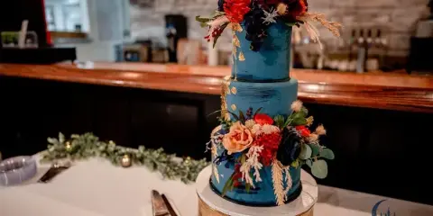 A three-tier blue wedding cake decorated with fresh flowers and gold accents on a table with a cake server.