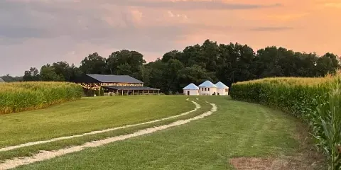 A wide shot of a farm venue at sunset, showing a large barn and three white grain silos in a grassy field bordered by corn.