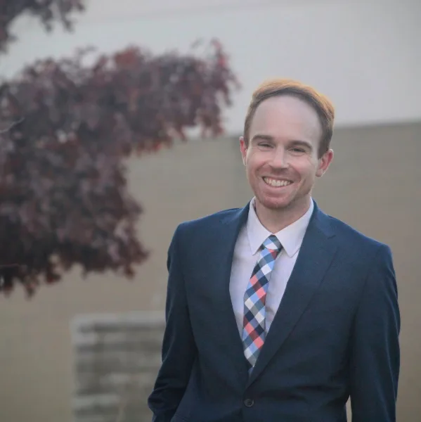 Portrait of a smiling man in a dark blue suit and patterned tie standing outdoors.