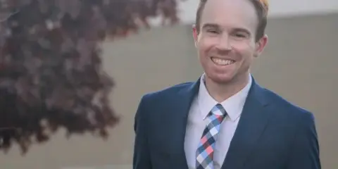 Portrait of a smiling man in a dark blue suit and patterned tie standing outdoors.