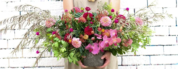 A person holding a large, lush floral arrangement with pink orchids, dahlias, and greenery against a white brick wall.