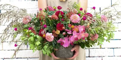 A person holding a large, lush floral arrangement with pink orchids, dahlias, and greenery against a white brick wall.