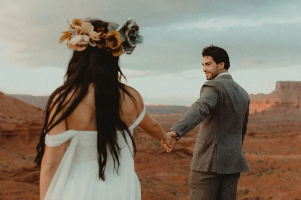A bride and groom holding hands in a desert landscape, with the bride wearing a floral crown and the groom in a grey suit.
