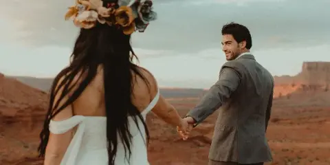 A bride and groom holding hands in a desert landscape, with the bride wearing a floral crown and the groom in a grey suit.