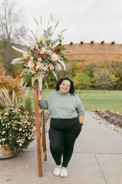 A woman standing next to a large, rustic floral arrangement on a wooden stand in an outdoor setting.