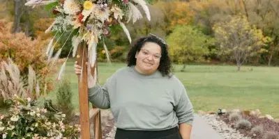 A woman standing next to a large, rustic floral arrangement on a wooden stand in an outdoor setting.